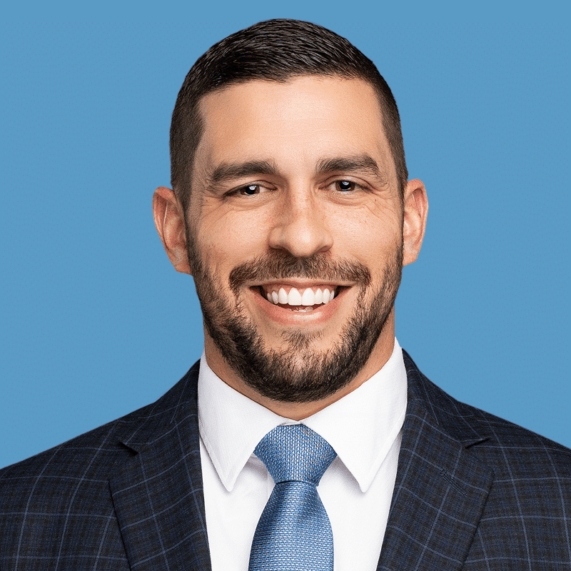 Headshot of Jared Baldwin, a smiling man with dark brown hair and a beard, wearing a navy-blue checkered suit and blue tie, set against a blue background.