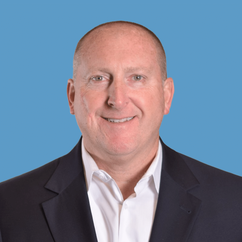 Headshot of Greg Blunt, a smiling bald man with a black suit and white button-down shirt, posed against a blue background.