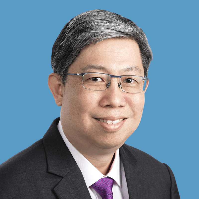 Headshot of Benedict Oon, a smiling Asian man with glasses wearing a suit and tie, posed against a blue background.
