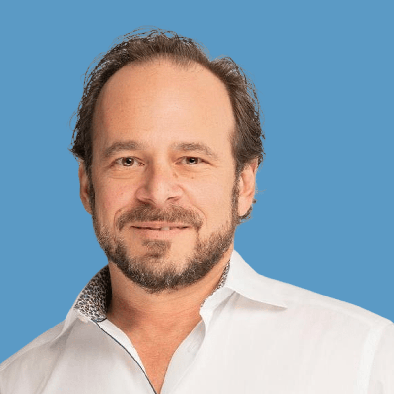 Headshot of Damien Breier, a middle-aged man with brown hair and a beard, wearing a white button-up shirt and smiling against a blue background.