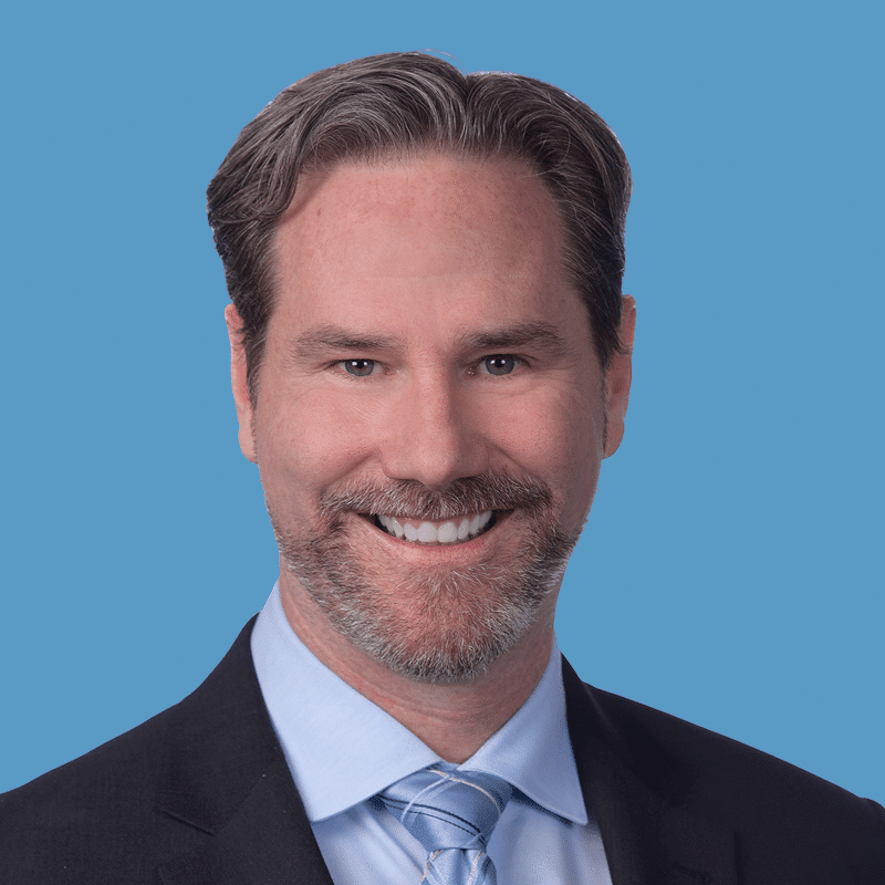Headshot of Rob Lucier, a smiling man with gray hair and beard wearing a suit and tie against a blue background.