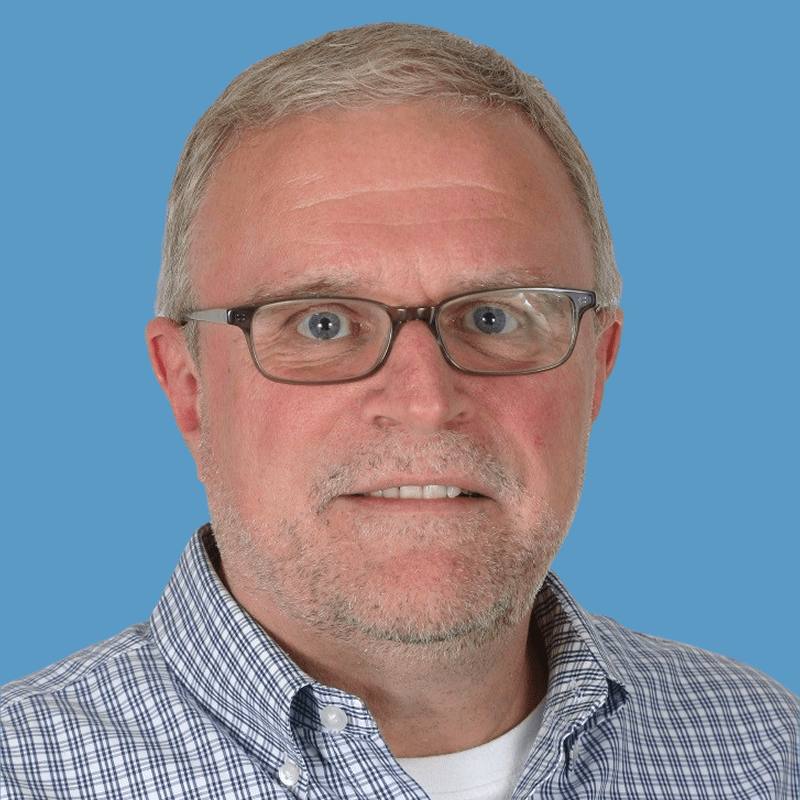 Headshot of William Roland, a smiling middle-aged man with gray hair and glasses wearing a plaid shirt against a blue background.