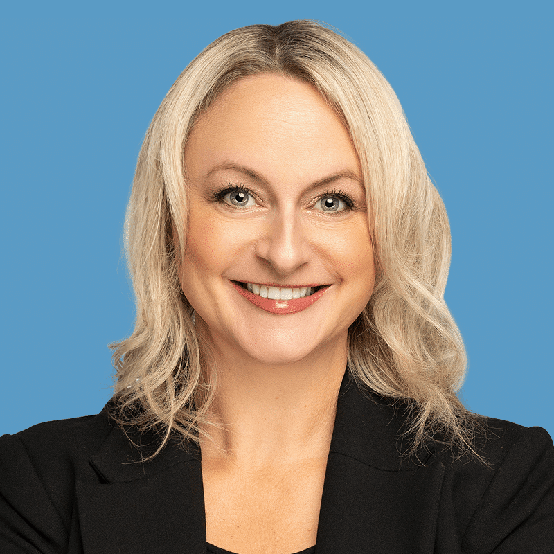 Headshot of Beth Abbott, a smiling woman with blonde hair wearing a black jacket against a blue background.