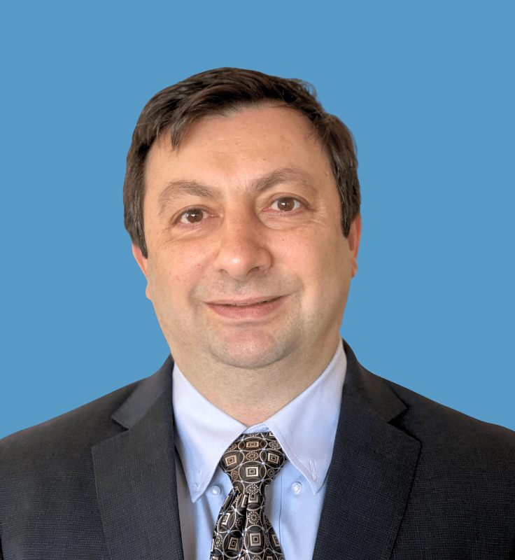 Headshot of Marius Nica, a smiling man wearing a dark suit jacket, light blue dress shirt, and patterned tie, posed against a solid blue background