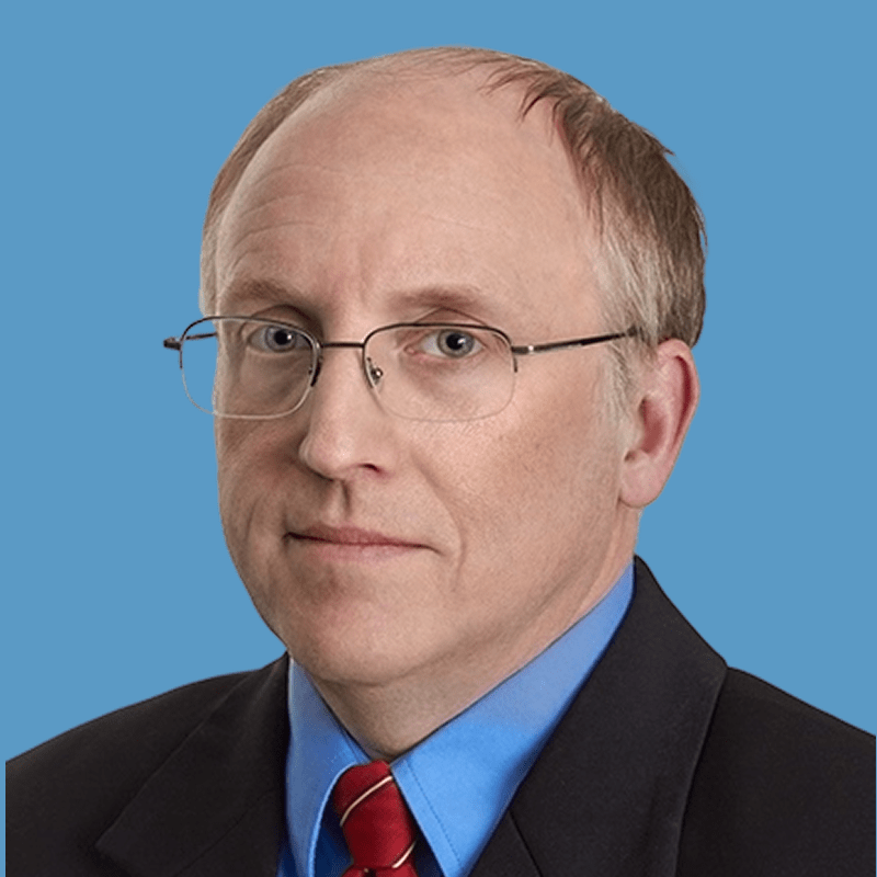 Headshot of Tony Lensing, a middle-aged man in formal business attire with glasses, sitting in front of blue background.