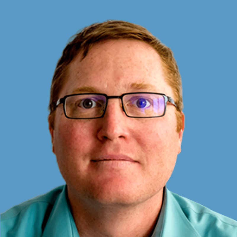 Headshot of John Whitehead, a smiling man wearing glasses and a green shirt against a blue background.