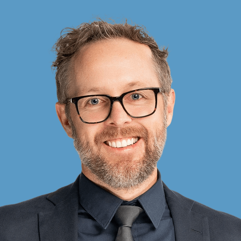 Headshot of Chris Wolvin, a smiling man with beard and glasses wearing a suit and tie against a blue background.